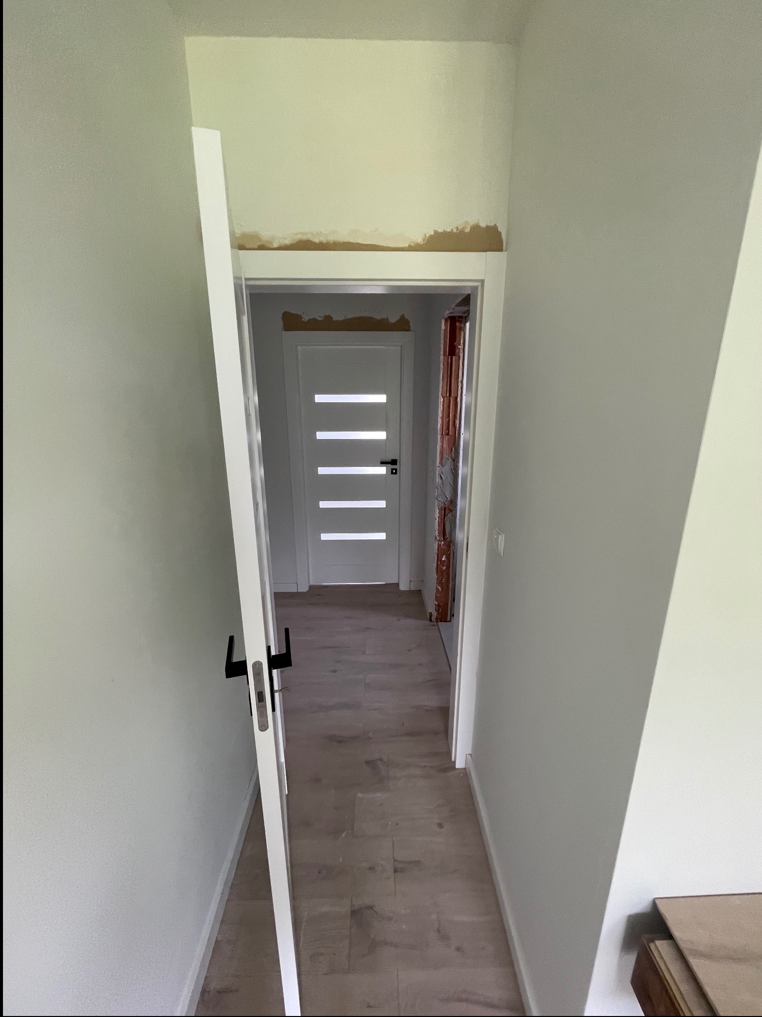 Wnętrze z częściowo zamontowanymi, białymi drzwiami z czarną klamką. Widoczne ślady po pracach wykończeniowych na ścianie nad ościeżnicą. Podłoga wyłożona panelami.