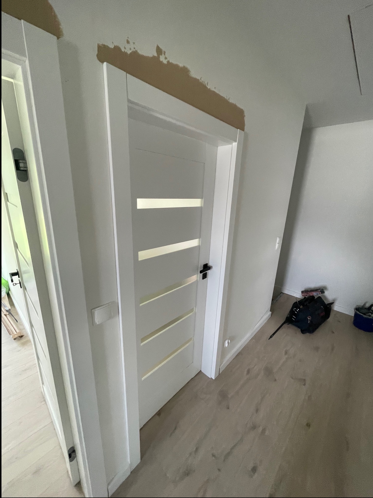 Białe drzwi z przeszkleniami w trakcie montażu, widoczne narzędzia i niedokończone wykończenie ściany nad ościeżnicą. Jasne wnętrze.