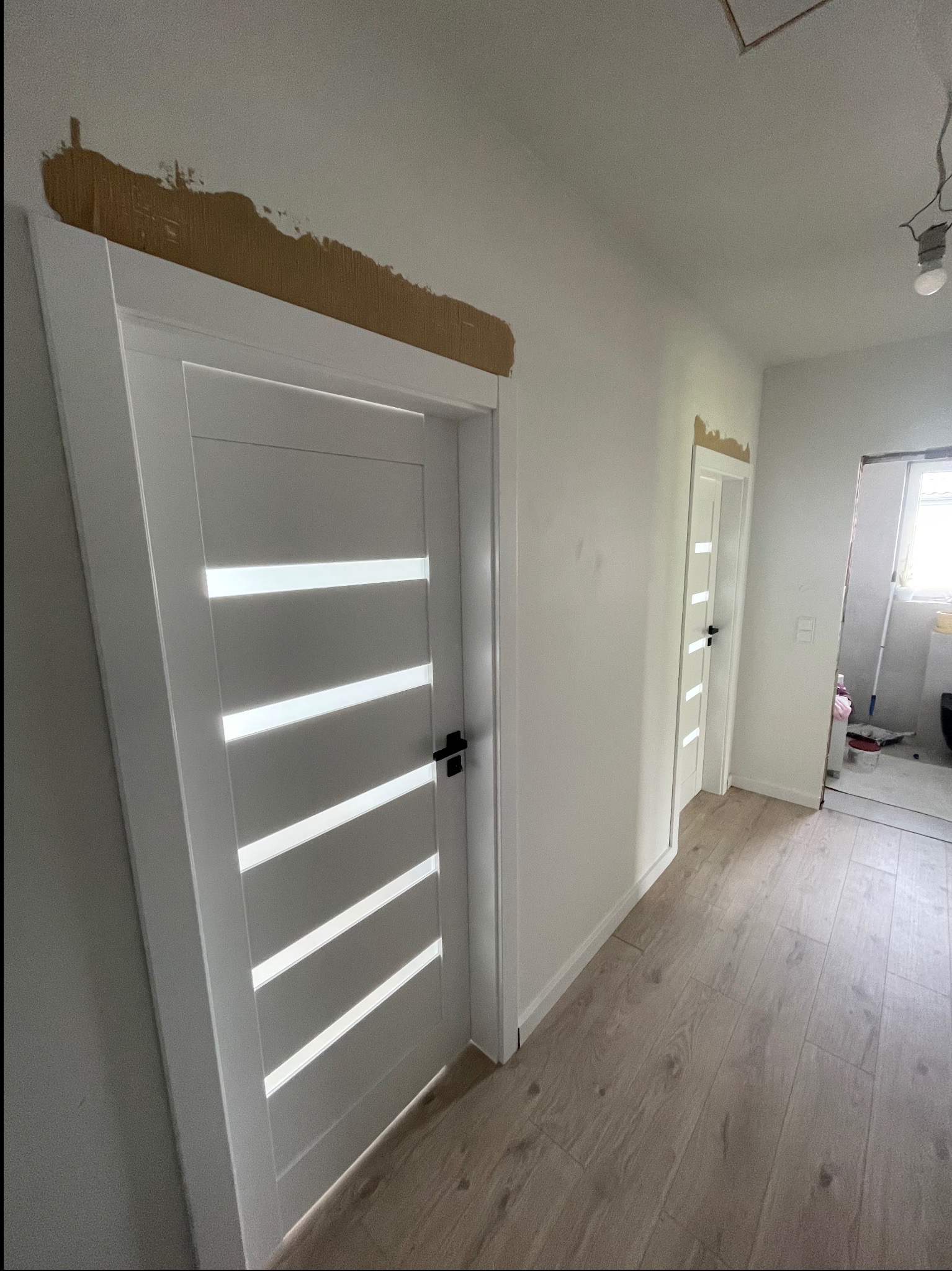 Białe, nowoczesne drzwi z poziomymi przeszkleniami w jasnym korytarzu. Widoczne ślady po remoncie na ścianie i podłodze z paneli. Kolejne drzwi w tle.