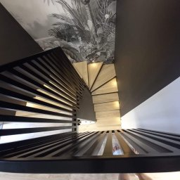 Robim wood - Spiralne schody z drewnianymi stopniami i nowoczesną, czarną balustradą z metalowych prętów. W tle ściana z motywem roślinnym.