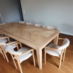 Długi, jasny stół z blatem w jodełkę otoczony krzesłami z jasną tapicerką, na drewnianej podłodze. Widok z góry.