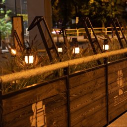 Robim wood - Rustykalna drewniana balustrada z liną i lampionami, oświetlona ciepłym światłem wieczornym, z logo 'Busola' na panelach. Dekoracja zewnętrzna.