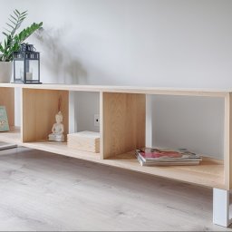 Robim wood - Jasna, drewniana szafka RTV z otwartymi półkami na białych, metalowych nogach. Na półkach książki, figurka Buddy i magazyny. Na górze roślina doniczkowa i lampion.