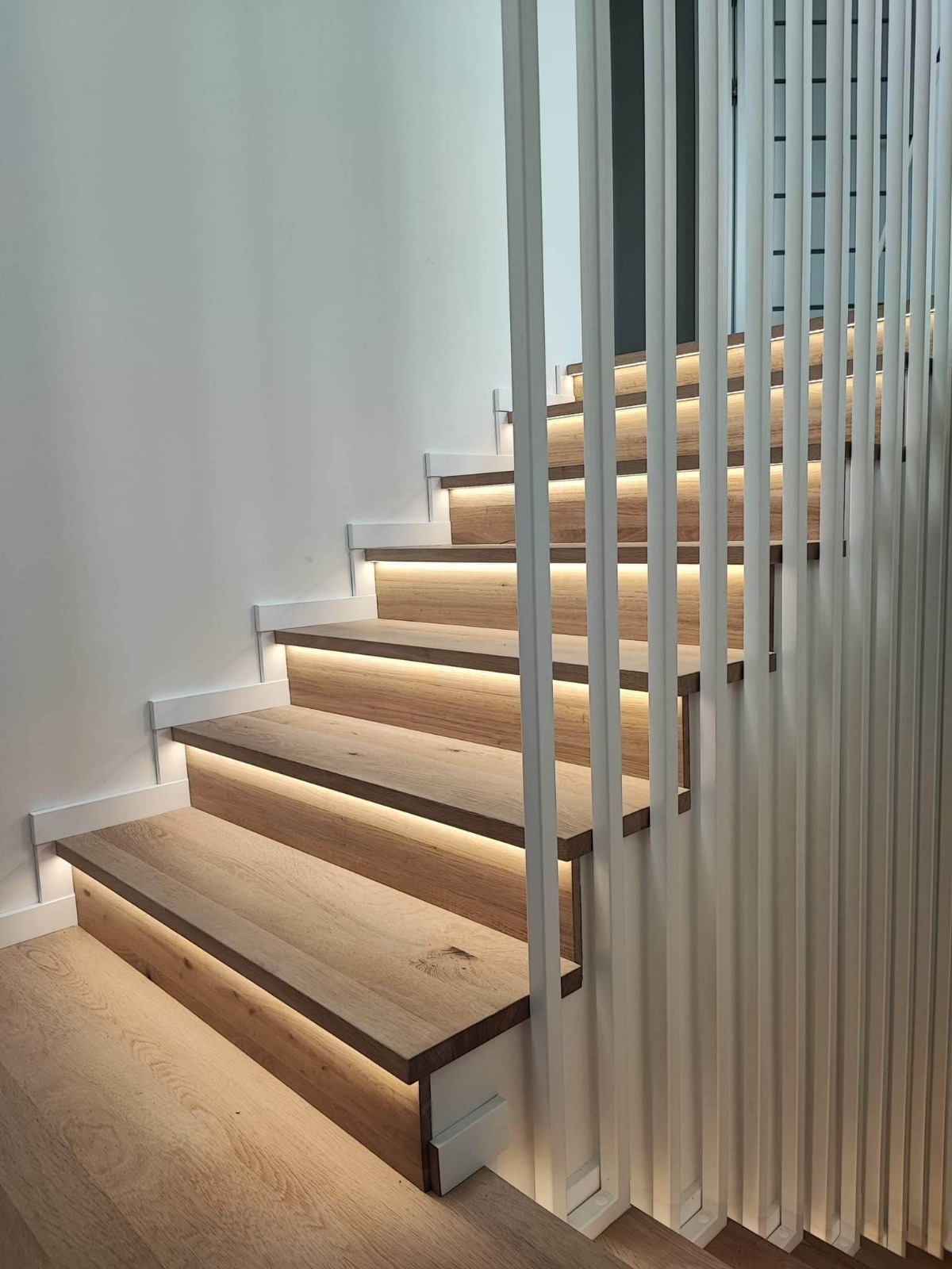 Nowoczesne schody drewniane z podświetleniem LED, białe barierki pionowe, jasne drewno, minimalistyczny design, widok z dołu na górę.