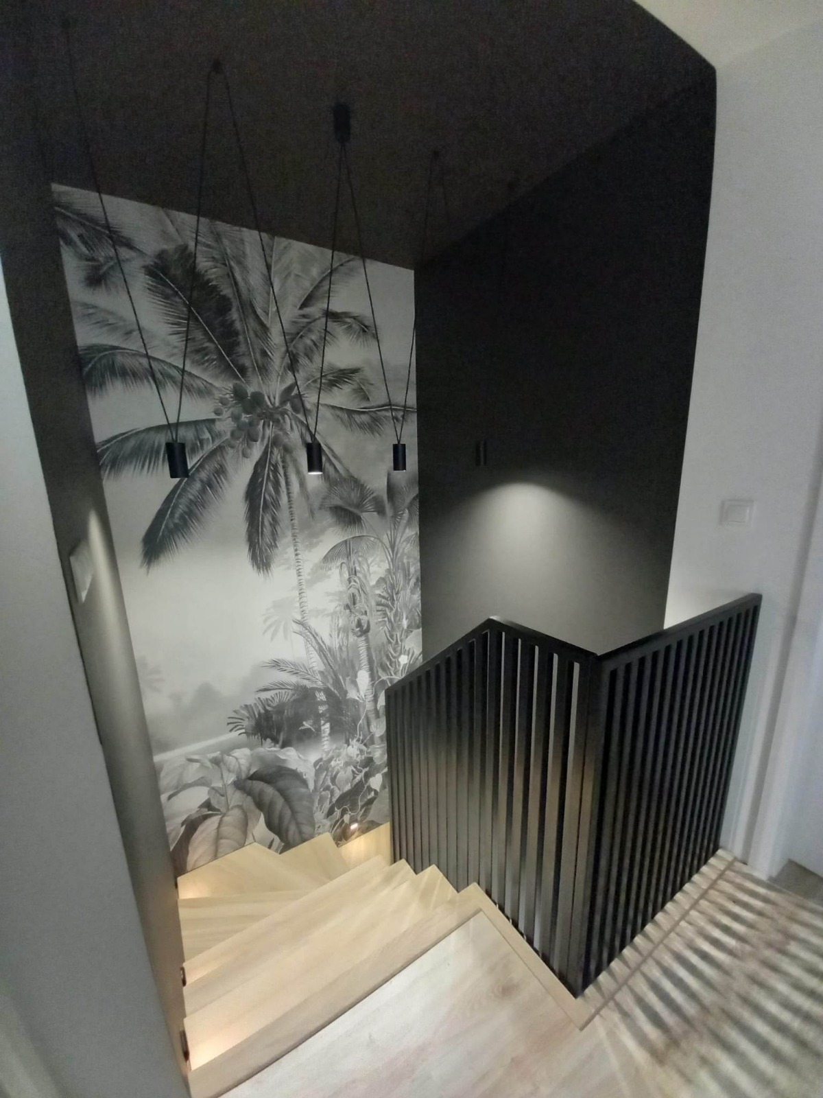 Jasne schody z nowoczesną, czarną balustradą na tle ściany z motywem tropikalnym i wiszącymi lampami. Minimalistyczny design, kontrast kolorów i faktur.