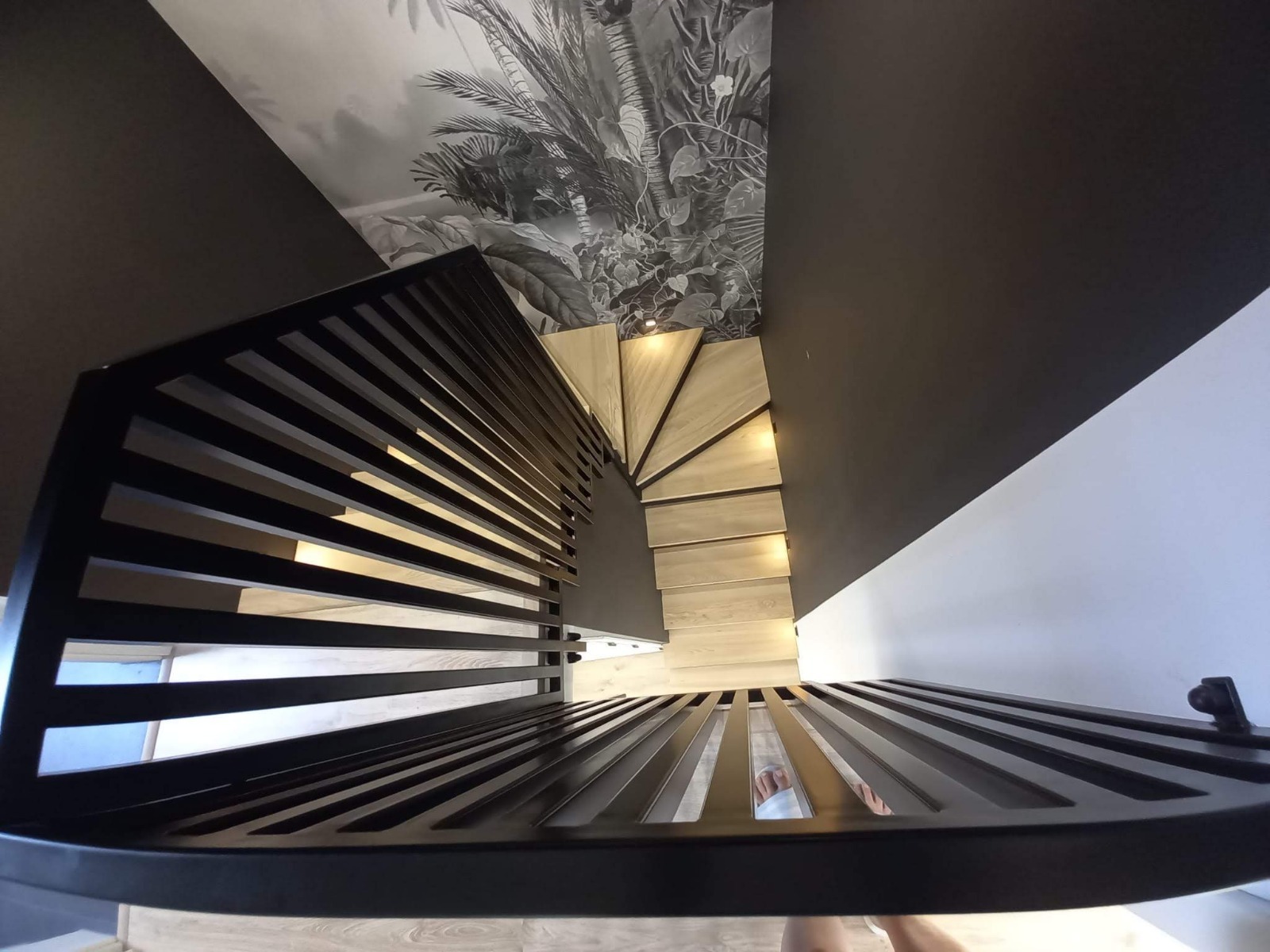 Spiralne schody z drewnianymi stopniami i nowoczesną, czarną balustradą z metalowych prętów. W tle ściana z motywem roślinnym.