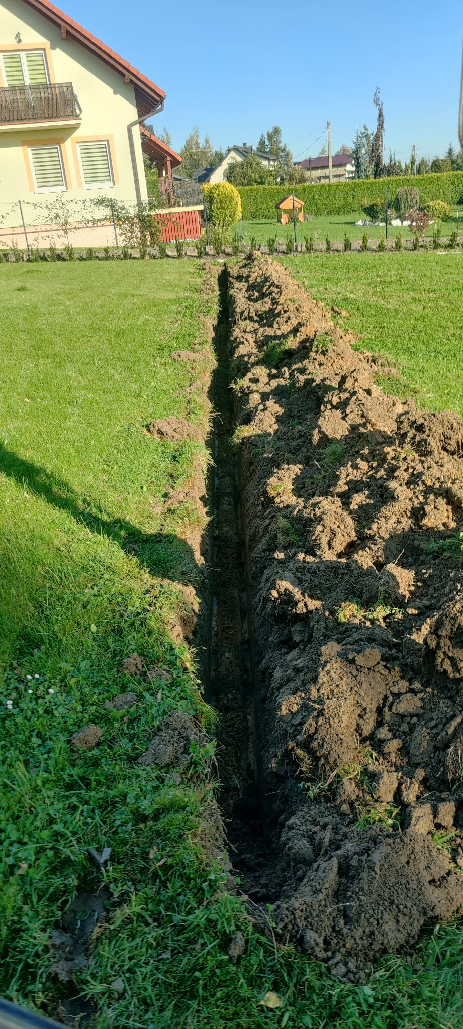 Wąski wykop w trawniku, prawdopodobnie pod instalacje, z widoczną ziemią po obu stronach rowu i domem w tle.