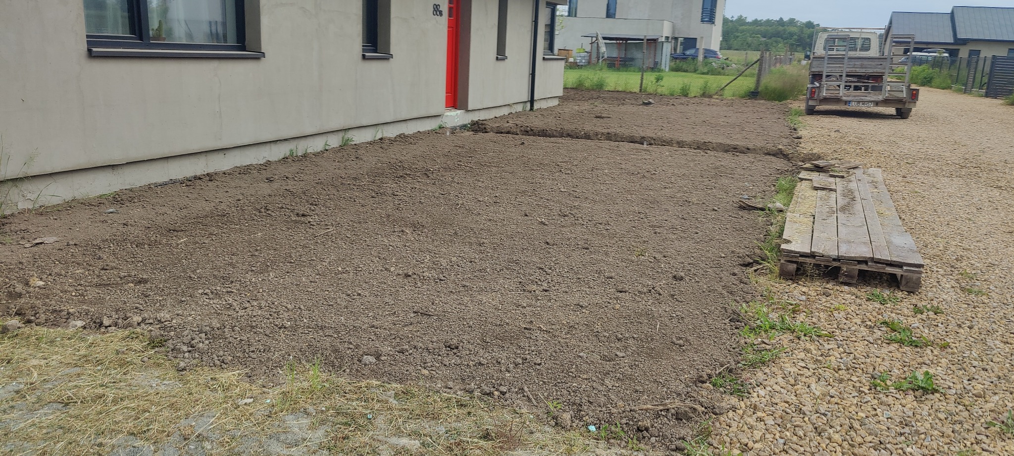 Przygotowany teren pod trawnik przy nowym domu z czerwoną stolarką drzwiową, widoczna świeżo nawieziona ziemia, wyrównana powierzchnia, w tle przyczepka samochodowa.