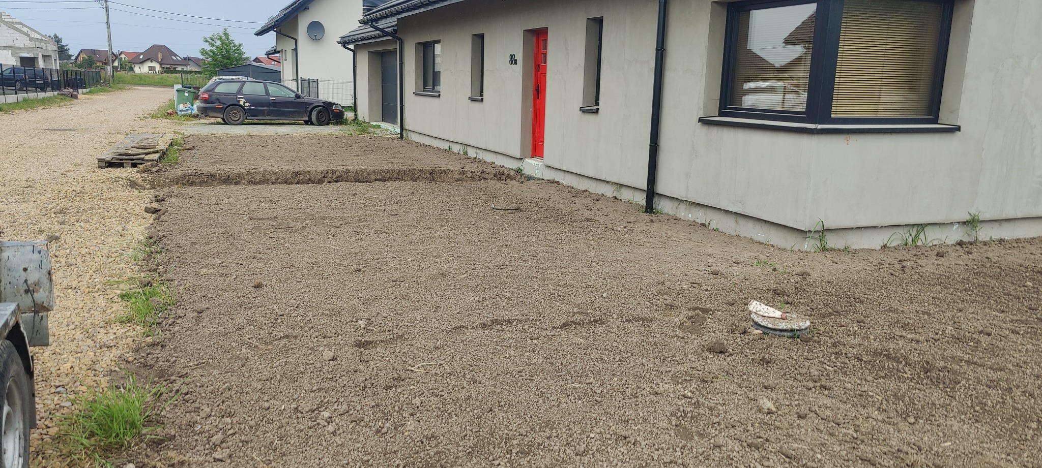 Przygotowany teren pod trawnik przy nowym domu, z wyrównaną ziemią i widocznymi śladami po maszynach, obok zaparkowany samochód osobowy i fragment przyczepki.