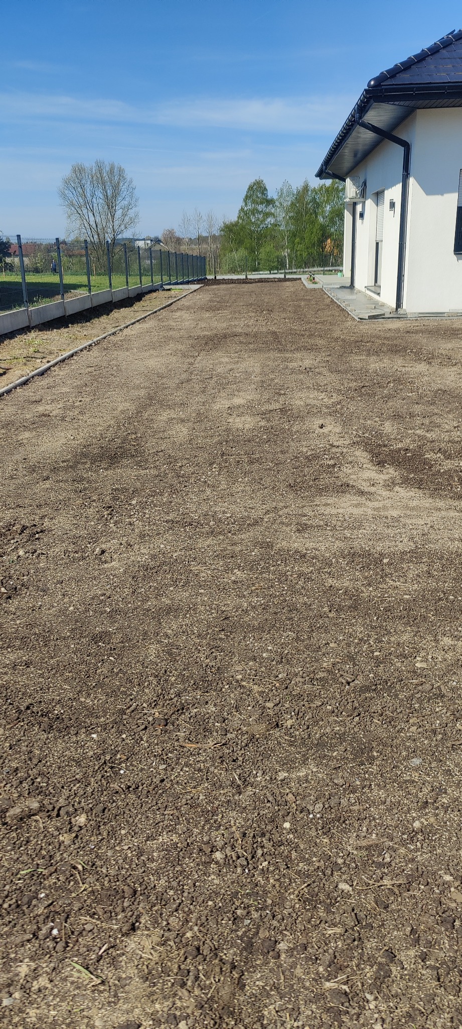 Przygotowany, wyrównany teren pod trawnik przy nowoczesnym domu jednorodzinnym z widoczną siatką ogrodzeniową w tle, w słoneczny dzień.