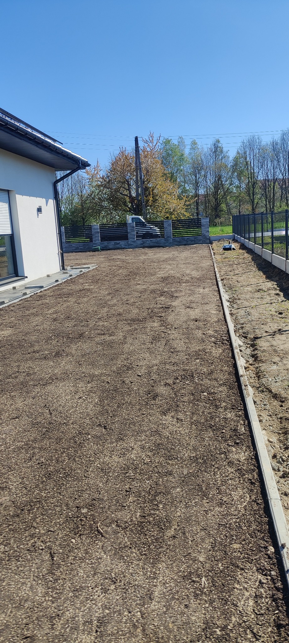 Uregulowany teren działki z przygotowaną podbudową pod trawnik, obrzeża betonowe oddzielają od sąsiedniej działki, w tle ogrodzenie panelowe i budynek mieszkalny.