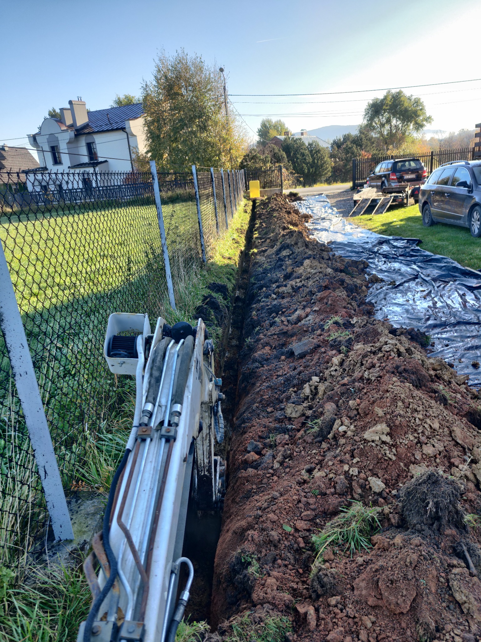 Wąski wykop w ziemi, wykonany przy użyciu minikoparki, zabezpieczony folią, z widocznymi samochodami i budynkiem w tle, w słoneczny dzień.