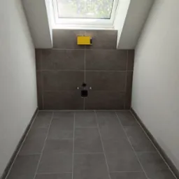 Przygotowana do montażu toalety łazienka na poddaszu z brązowymi płytkami podłogowymi i ściennymi, widoczne wyprowadzenia wodne i kanalizacyjne oraz okno dachowe.