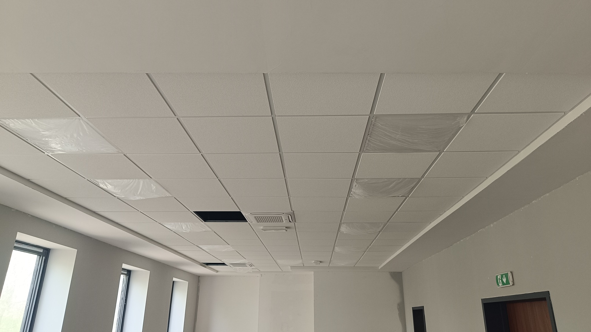 Montaż sufitu podwieszanego kasetonowego w biurze, częściowo zabezpieczony folią, widoczne otwory na klimatyzację i okna w tle.