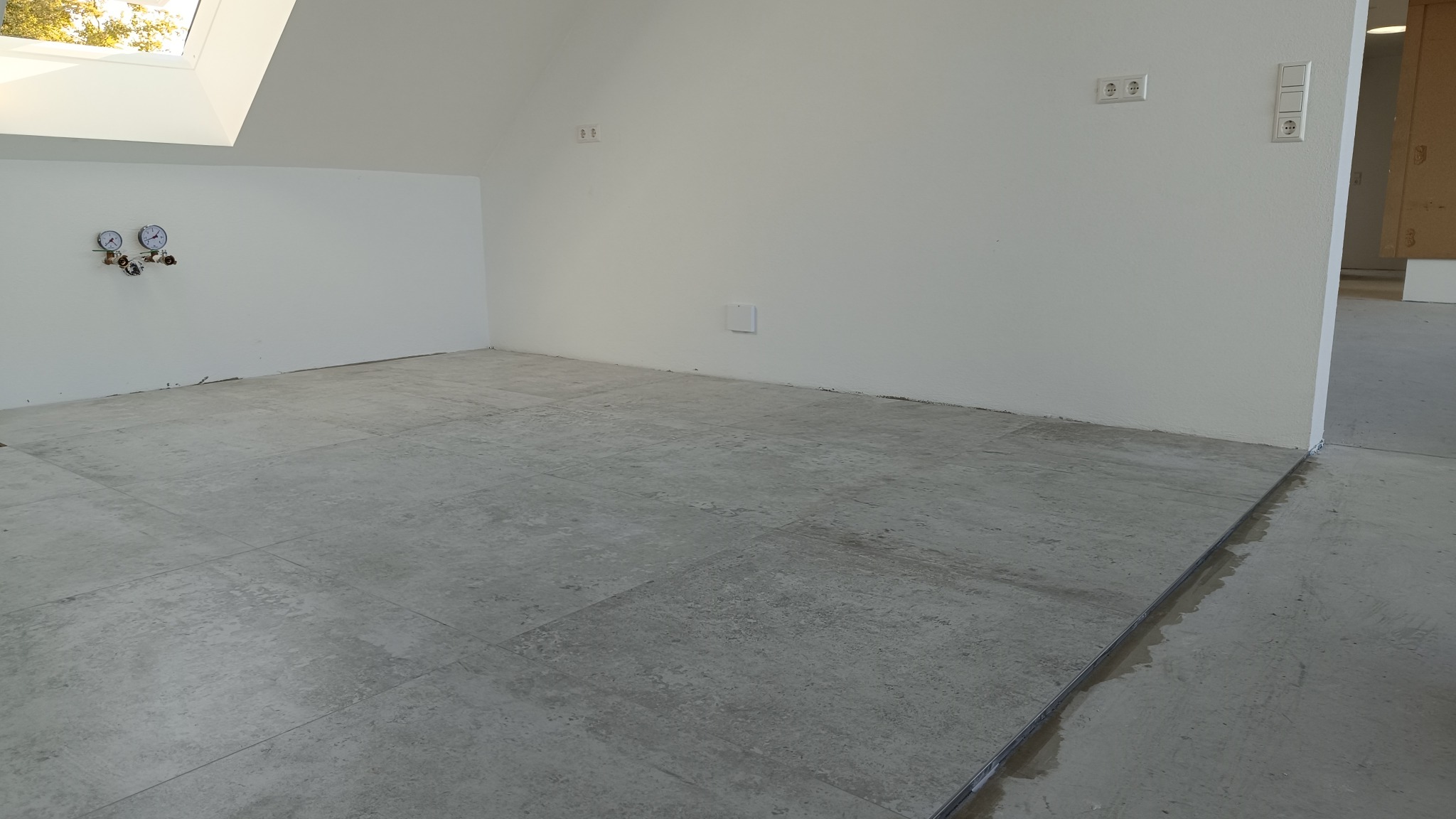 Szare płytki podłogowe ułożone w jasnym pomieszczeniu na poddaszu ze świetlikiem dachowym, widoczne instalacje na ścianie i gniazdka elektryczne.