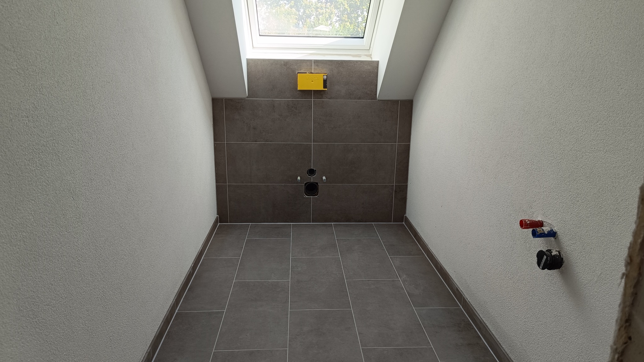 Przygotowana do montażu toalety łazienka na poddaszu z brązowymi płytkami podłogowymi i ściennymi, widoczne wyprowadzenia wodne i kanalizacyjne oraz okno dachowe.