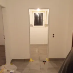 Wnętrze w trakcie malowania: widoczne świeżo pomalowane ściany, zabezpieczone taśmą malarską framugi drzwi i okien, puszka farby na folii ochronnej oraz szpachelka na podłodze z płytek.