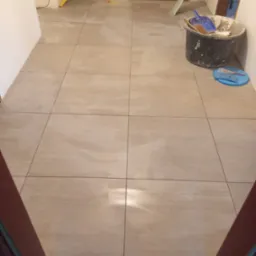 Beżowe płytki podłogowe ułożone w korytarzu, widoczny kubeł z narzędziami glazurniczymi i poziomica oparta o ścianę.