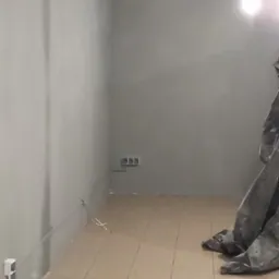 Pomieszczenie w trakcie remontu, szare ściany, brązowa podłoga z płytek, zabezpieczona folią malarską część podłogi, widoczne gniazdka elektryczne i wystające kable.