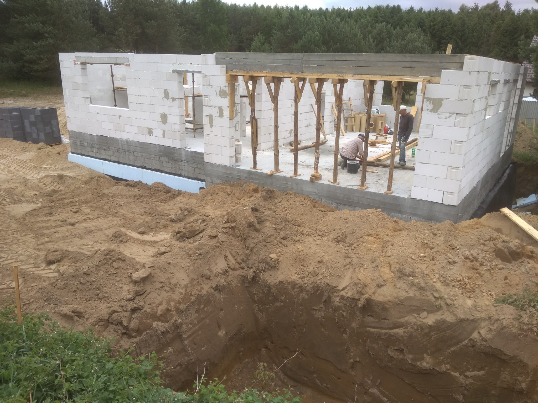 Budowa domu z białych bloczków: widok na fundament z izolacją, ściany w trakcie wznoszenia, drewniane podpory stropu i dwóch pracowników wewnątrz konstrukcji.