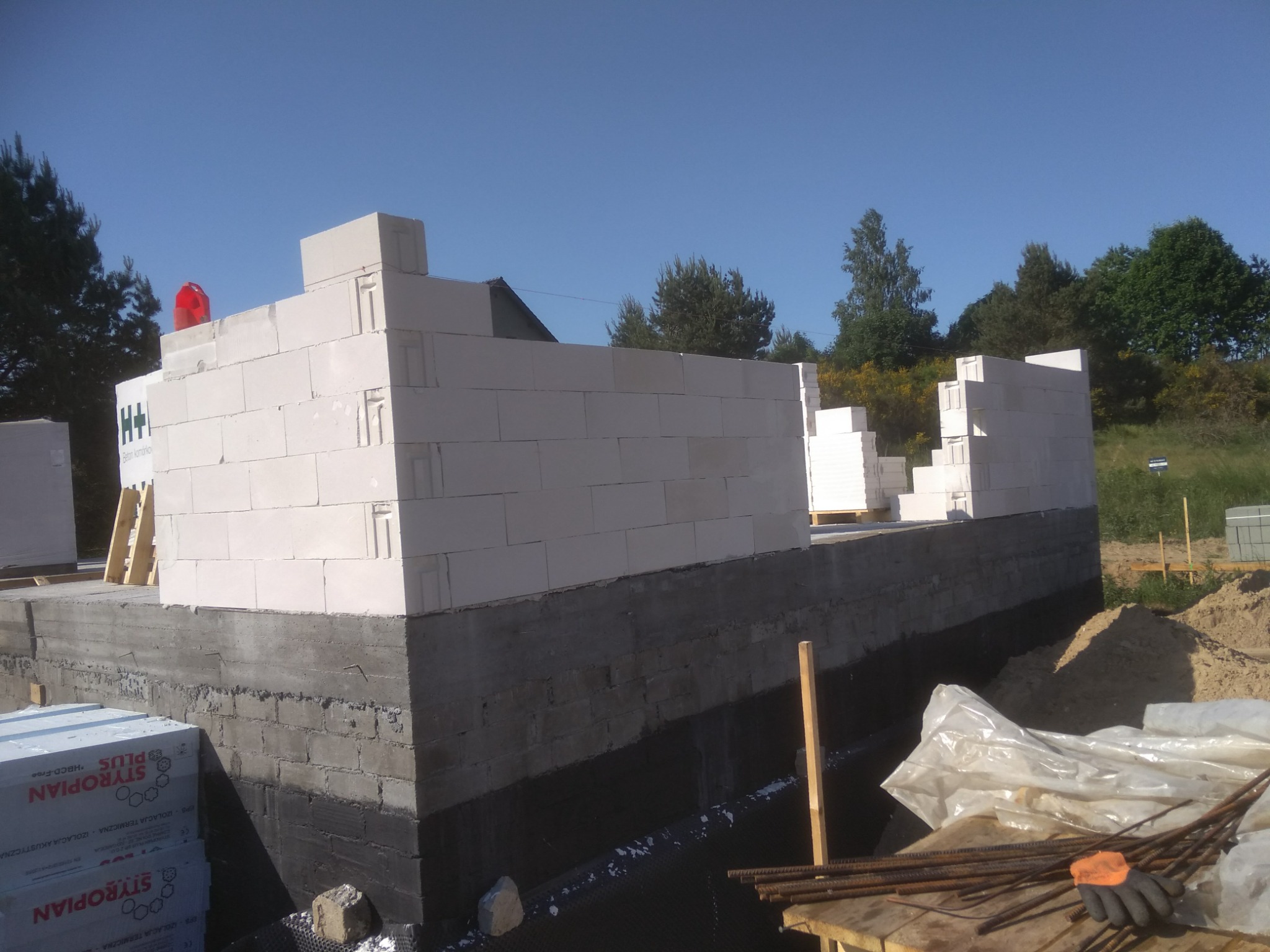 Wznoszenie ścian parteru z białych bloczków na fundamencie z izolacją przeciwwilgociową, widoczne materiały budowlane i narzędzia na placu budowy, w tle zielone drzewa i błękitne niebo.
