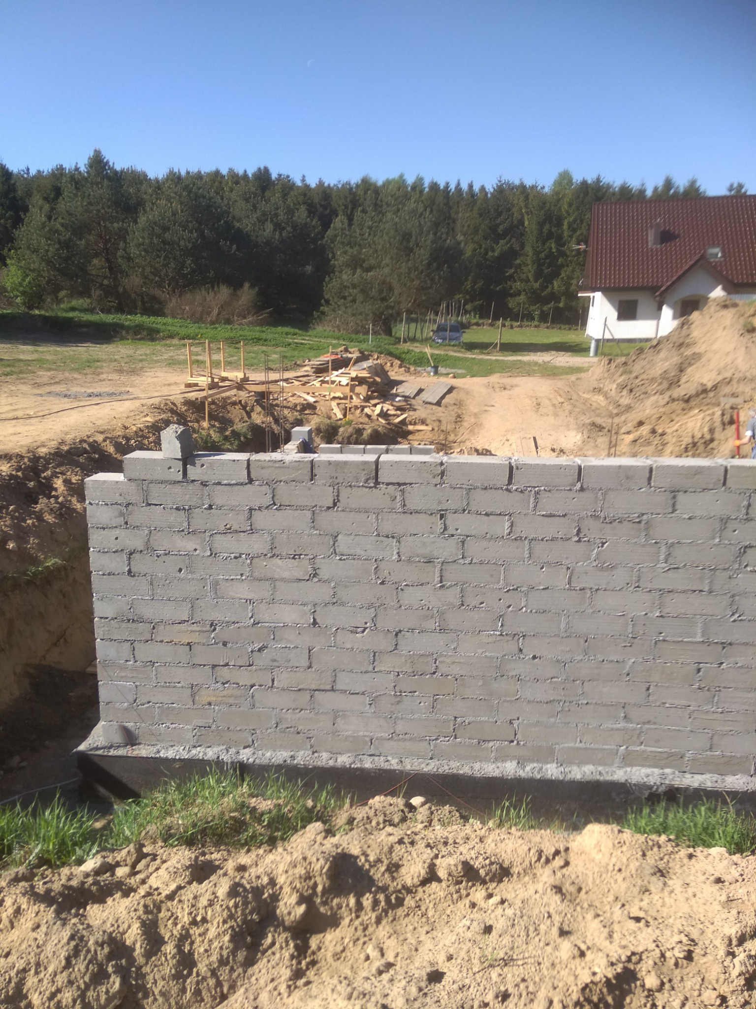 Wznoszenie fundamentów z bloczków betonowych na placu budowy, widoczne wykopane fundamenty, w tle las i dom w stanie surowym otwartym.