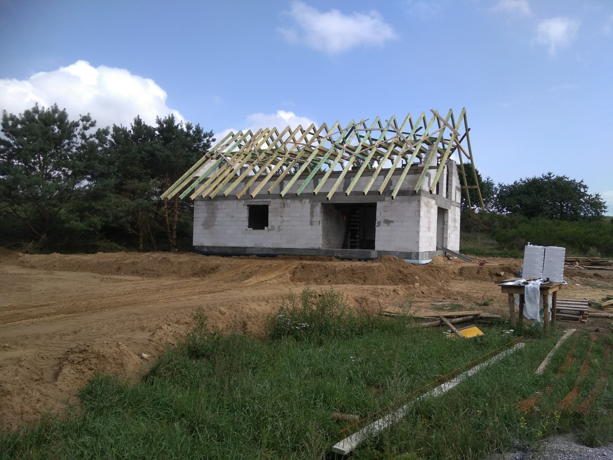 Surowy stan domu murowanego z pustaków, w trakcie budowy dachu, z widocznymi więźbami dachowymi zaimpregnowanymi na zielono, na tle nieba z chmurami i drzew