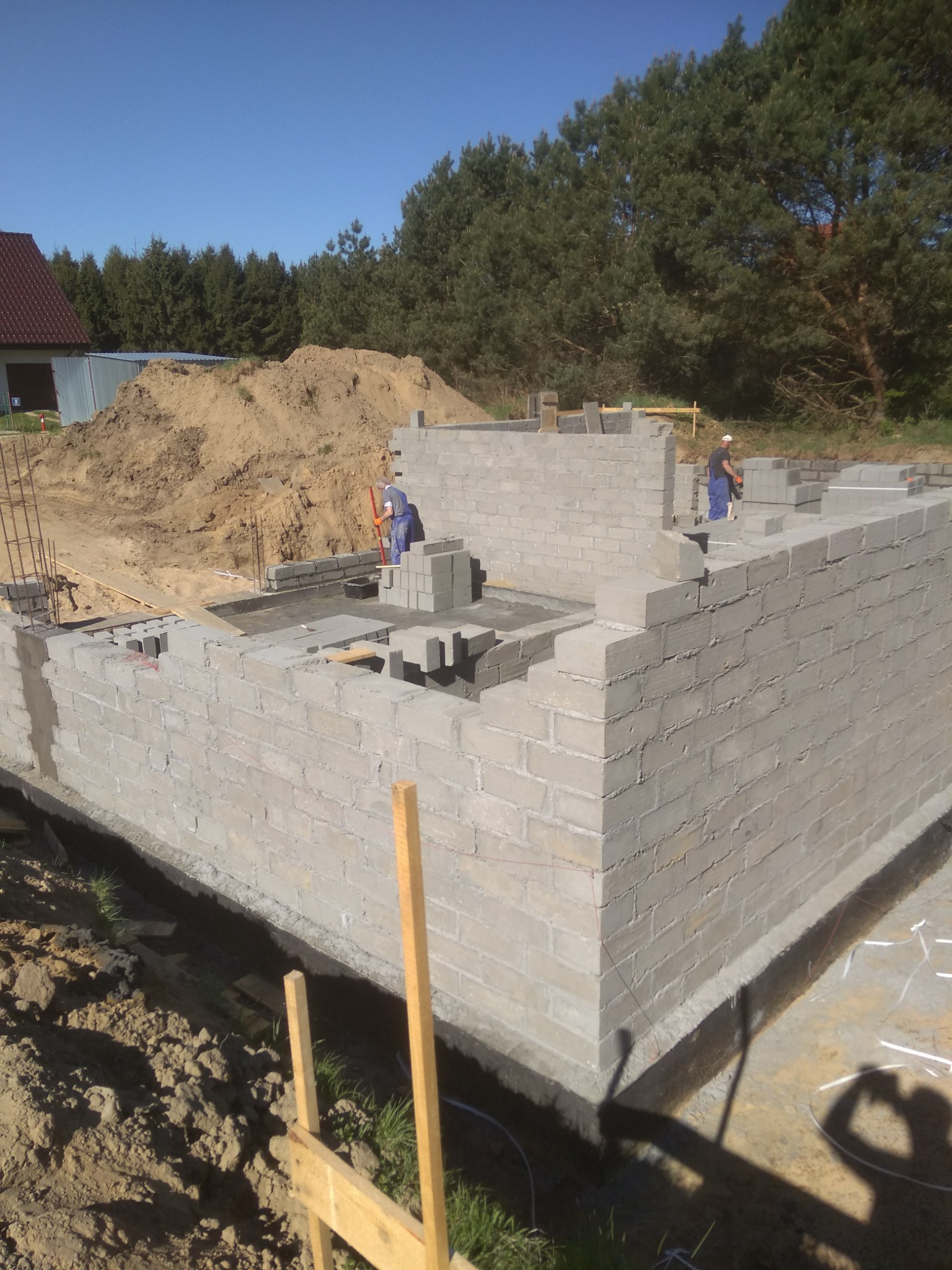 Budowa fundamentów z bloczków betonowych na tle lasu, dwóch robotników w niebieskich kombinezonach murowujących ściany, widoczna sterta piasku i częściowo wybudowany dom w oddali.