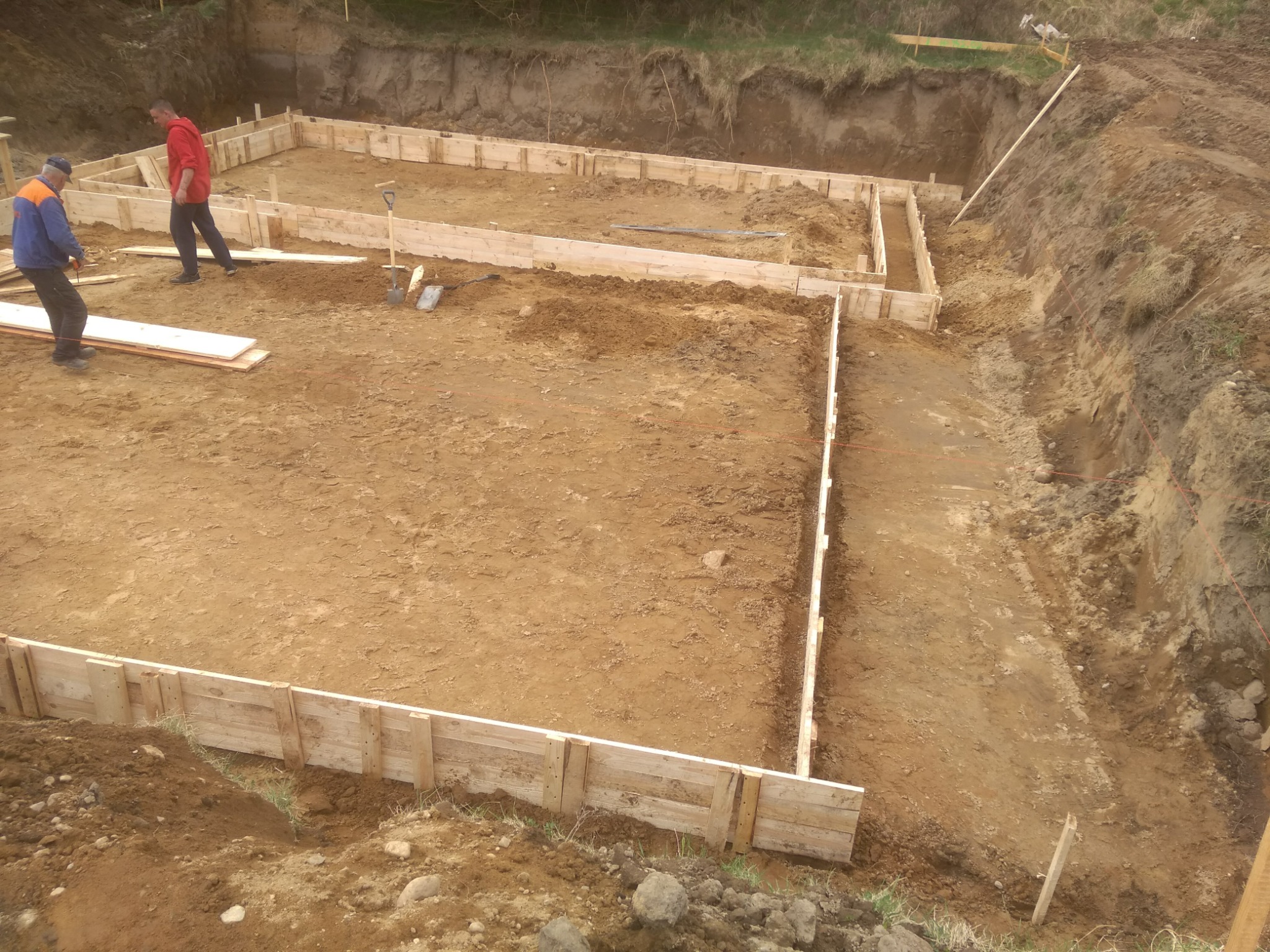 Przygotowanie fundamentów pod budowę domu: drewniany szalunek tworzy ramy, widoczny wykop i dwóch pracowników budowlanych.