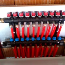 Rozdzielacz ogrzewania podłogowego w szafce z licznymi czerwonymi rurami, zawory z niebieskimi i czerwonymi pokrętłami, widoczne mosiężne złączki.