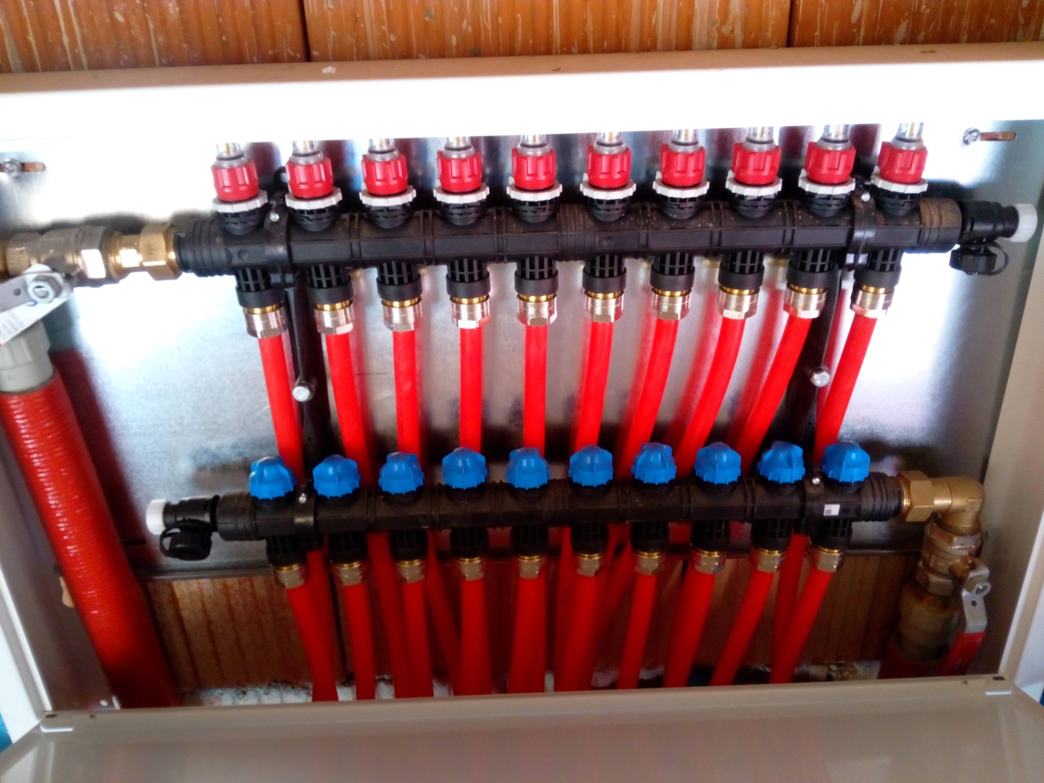 Rozdzielacz ogrzewania podłogowego w szafce z licznymi czerwonymi rurami, zawory z niebieskimi i czerwonymi pokrętłami, widoczne mosiężne złączki.