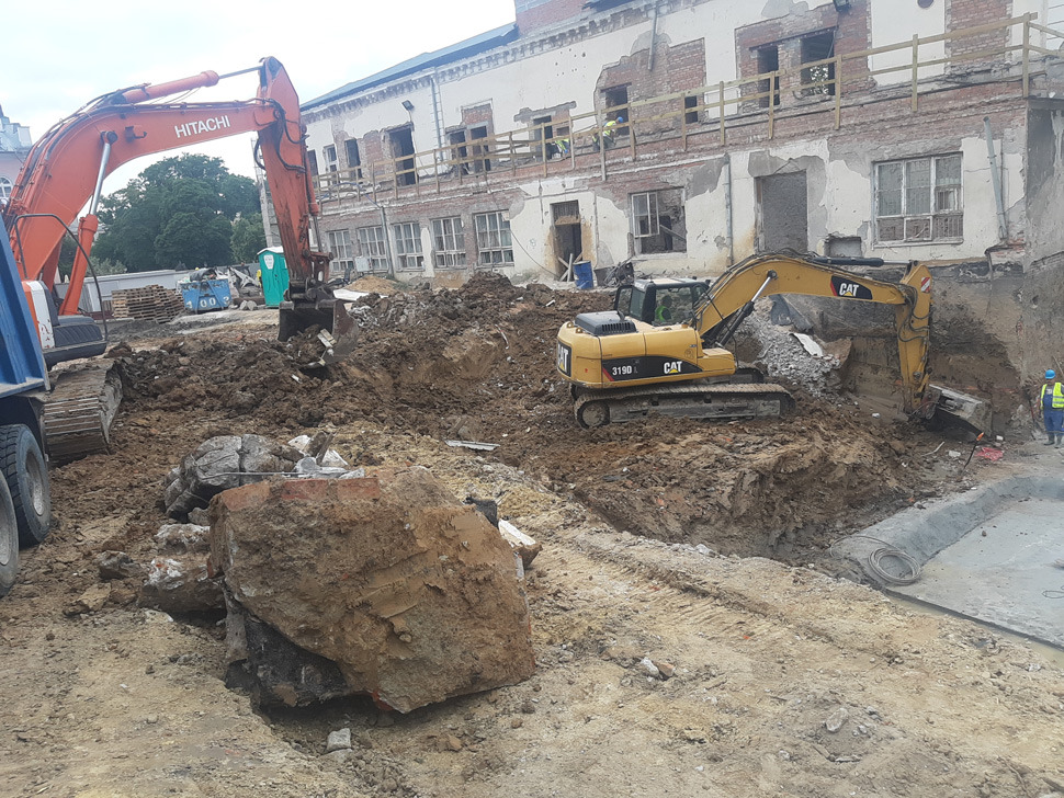 Dwie koparki, Hitachi i CAT 319D, pracują na placu budowy z częściowo rozebranym budynkiem w tle. W wykopie widoczne fragmenty gruzu i ziemi, a obok betonowa konstrukcja.