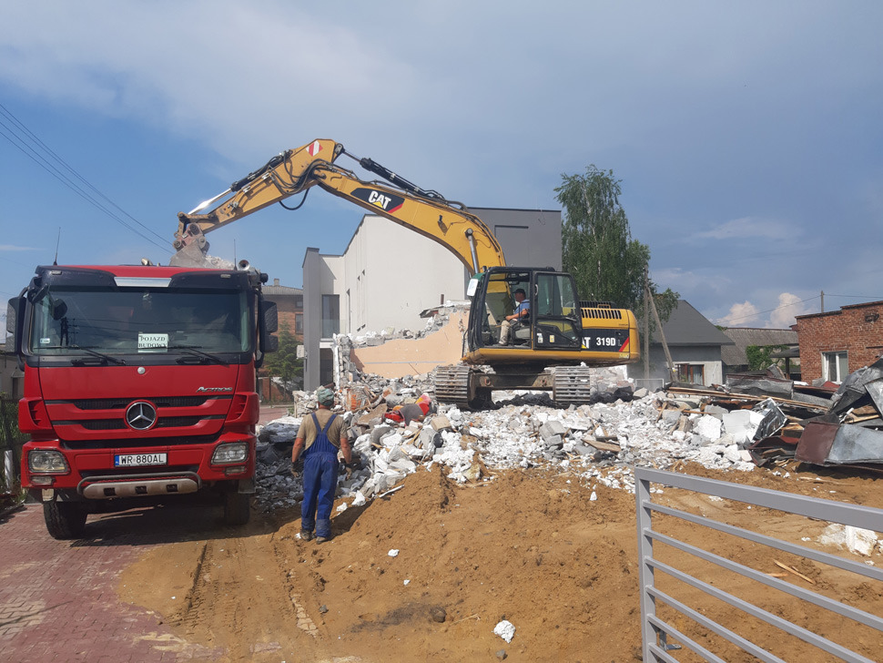 Czerwona ciężarówka Mercedes-Benz i żółta koparka CAT 319D na placu budowy podczas rozbiórki, z pracownikiem w kombinezonie na pierwszym planie i stertą gruzu oraz ziemi.