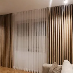 Salon z panelowymi, beżowymi i białymi, pionowymi zasłonami, widok na fragment sofy i stołu z krzesłami.