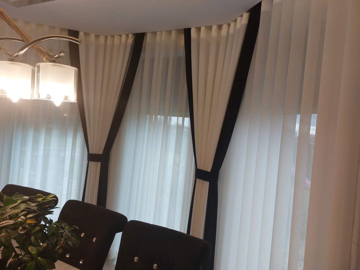 Eleganckie, długie, białe zasłony z czarnymi obramowaniami, upięte paskami, widoczne przy stole z krzesłami i lampą
