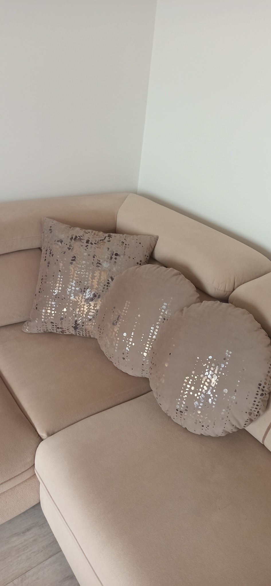 Beżowa sofa narożna z kwadratową i dwiema okrągłymi poduszkami dekoracyjnymi z błyszczącymi, srebrnymi aplikacjami.