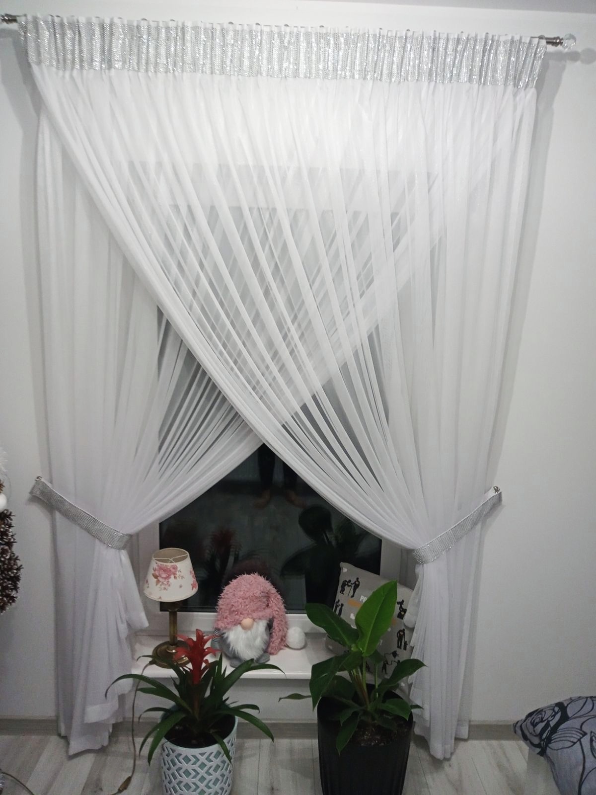 Delikatne, białe zasłony woalowe z srebrnymi, błyszczącymi zdobieniami na karniszu i upięciach, wiszące w oknie z widocznymi roślinami doniczkowymi i lampką na parapecie.