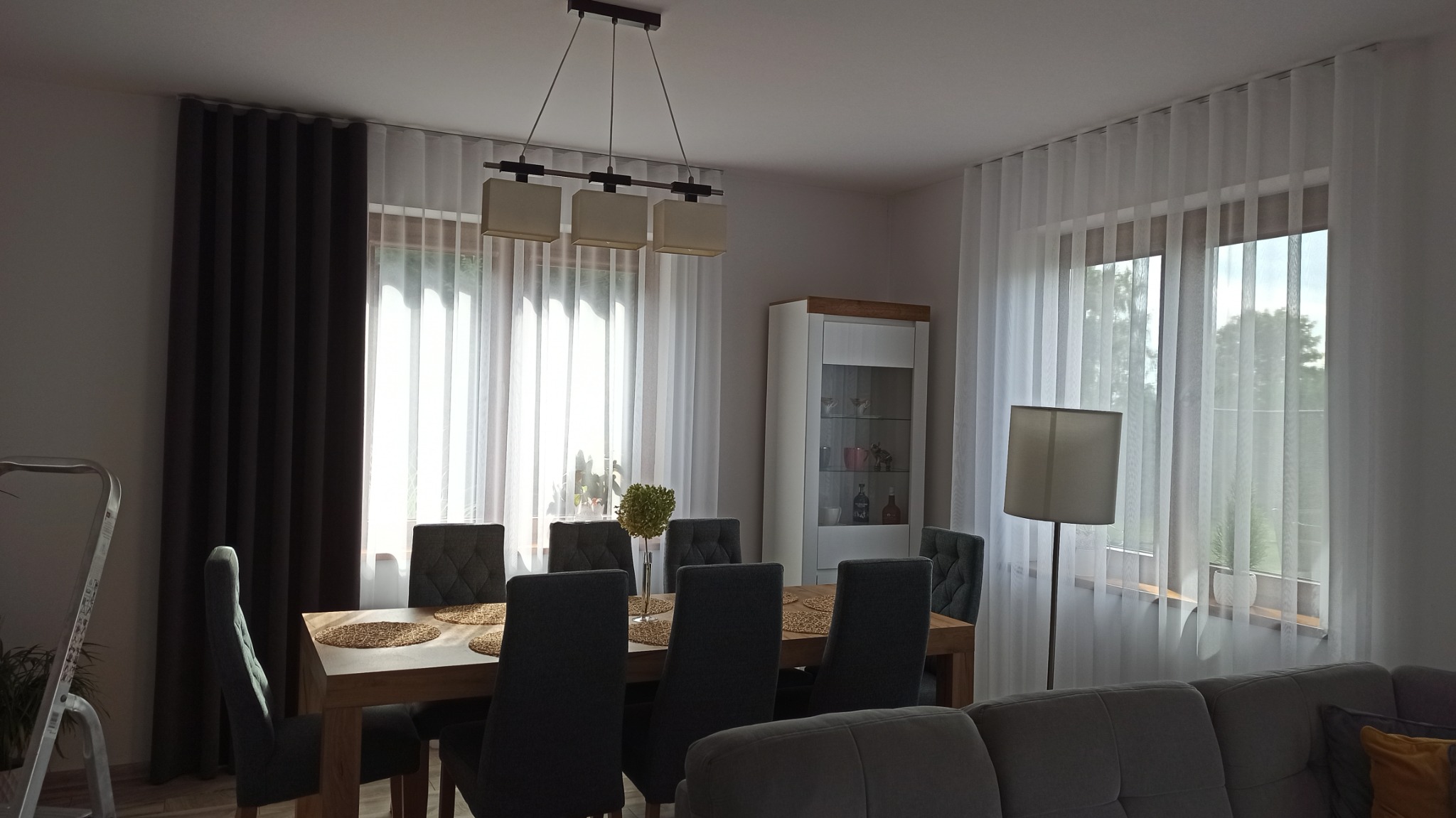 Jadalnia z drewnianym stołem i sześcioma krzesłami, okno przesłonięte białymi, delikatnymi firanami i ciemnoszarymi, grubymi zasłonami, lampa wisząca nad stołem.