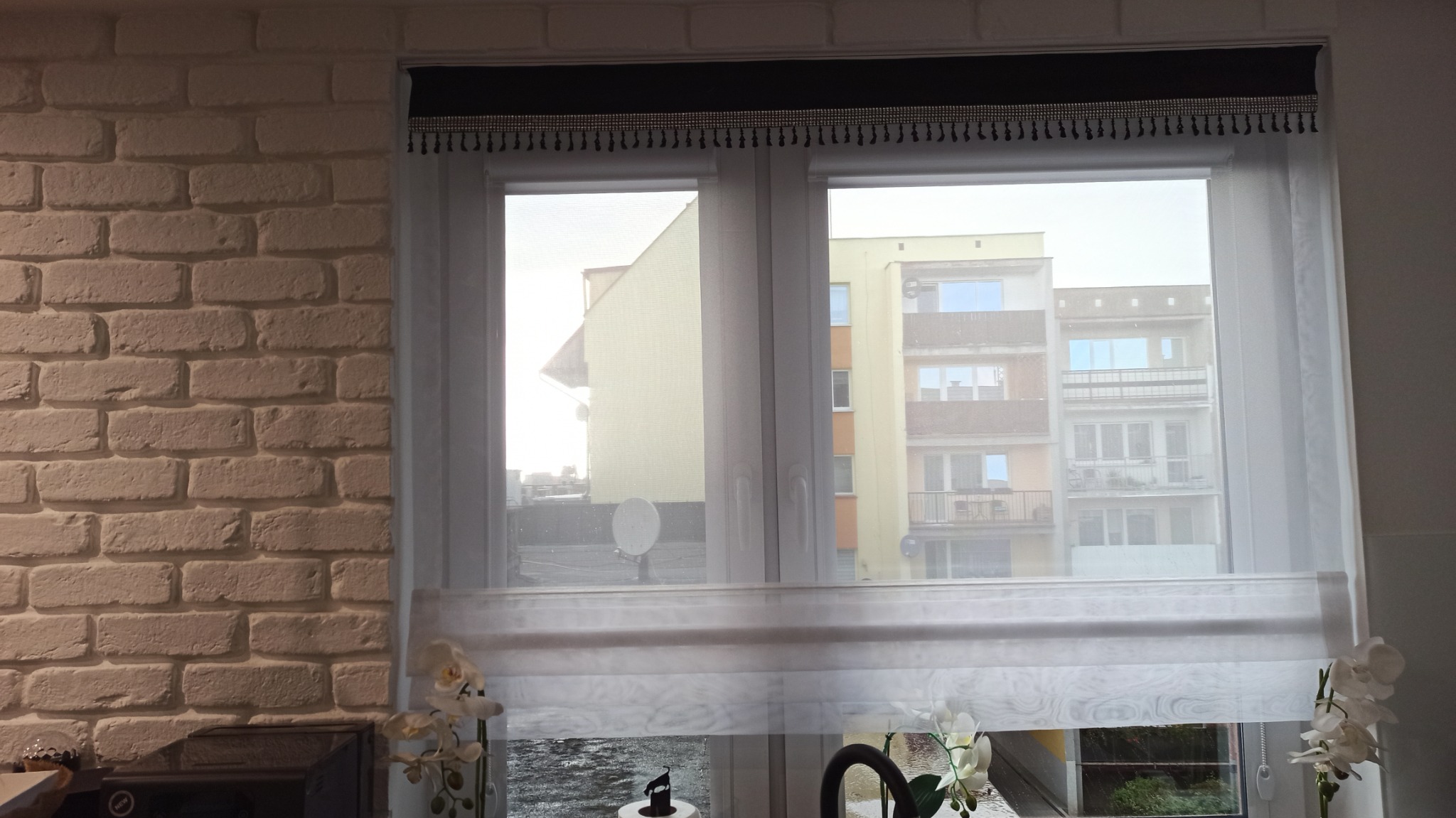 Delikatne, białe zasłony panelowe z czarnym, zdobionym górnym pasem, widoczne na tle okna z widokiem na budynki w Ząbrowie, obok dekoracyjne kwiaty.