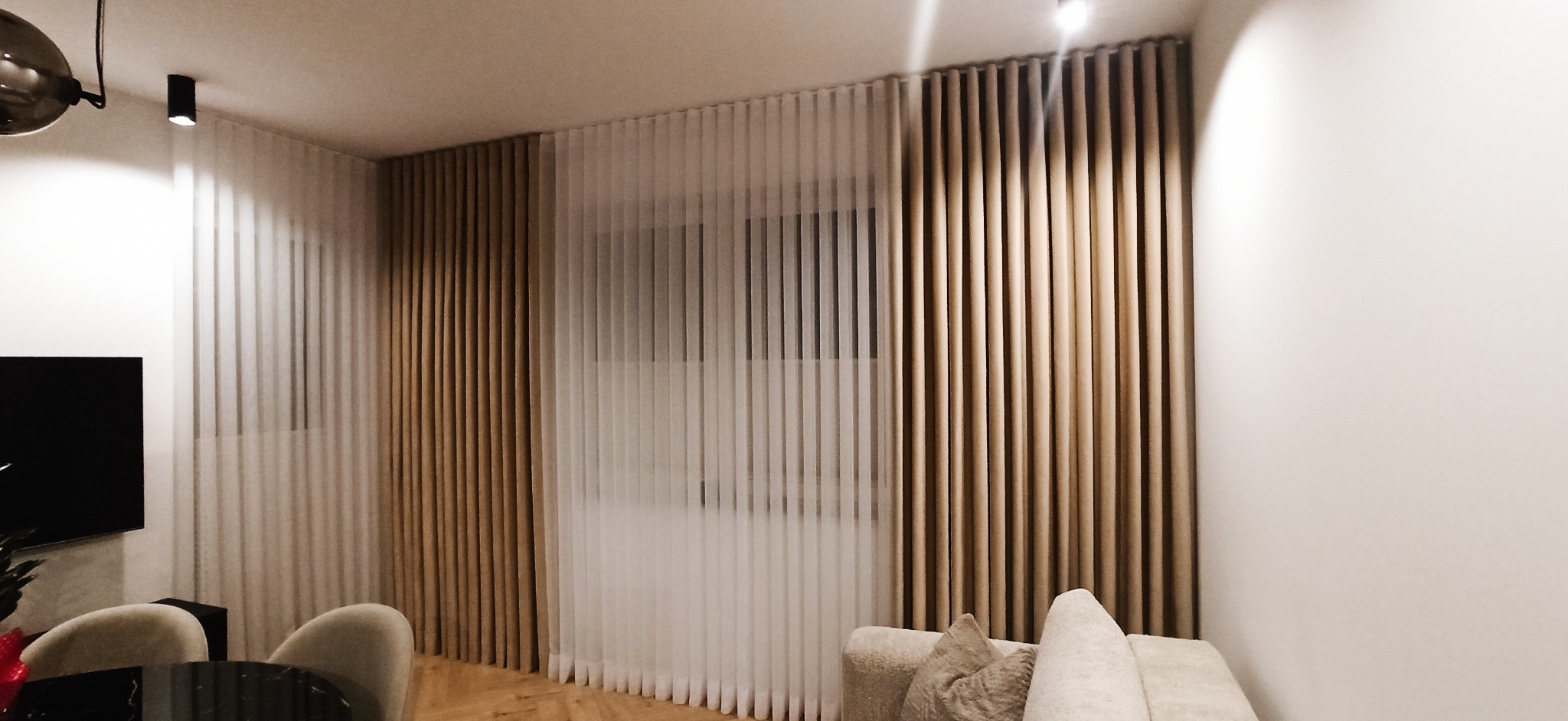 Salon z panelowymi, beżowymi i białymi, pionowymi zasłonami, widok na fragment sofy i stołu z krzesłami.