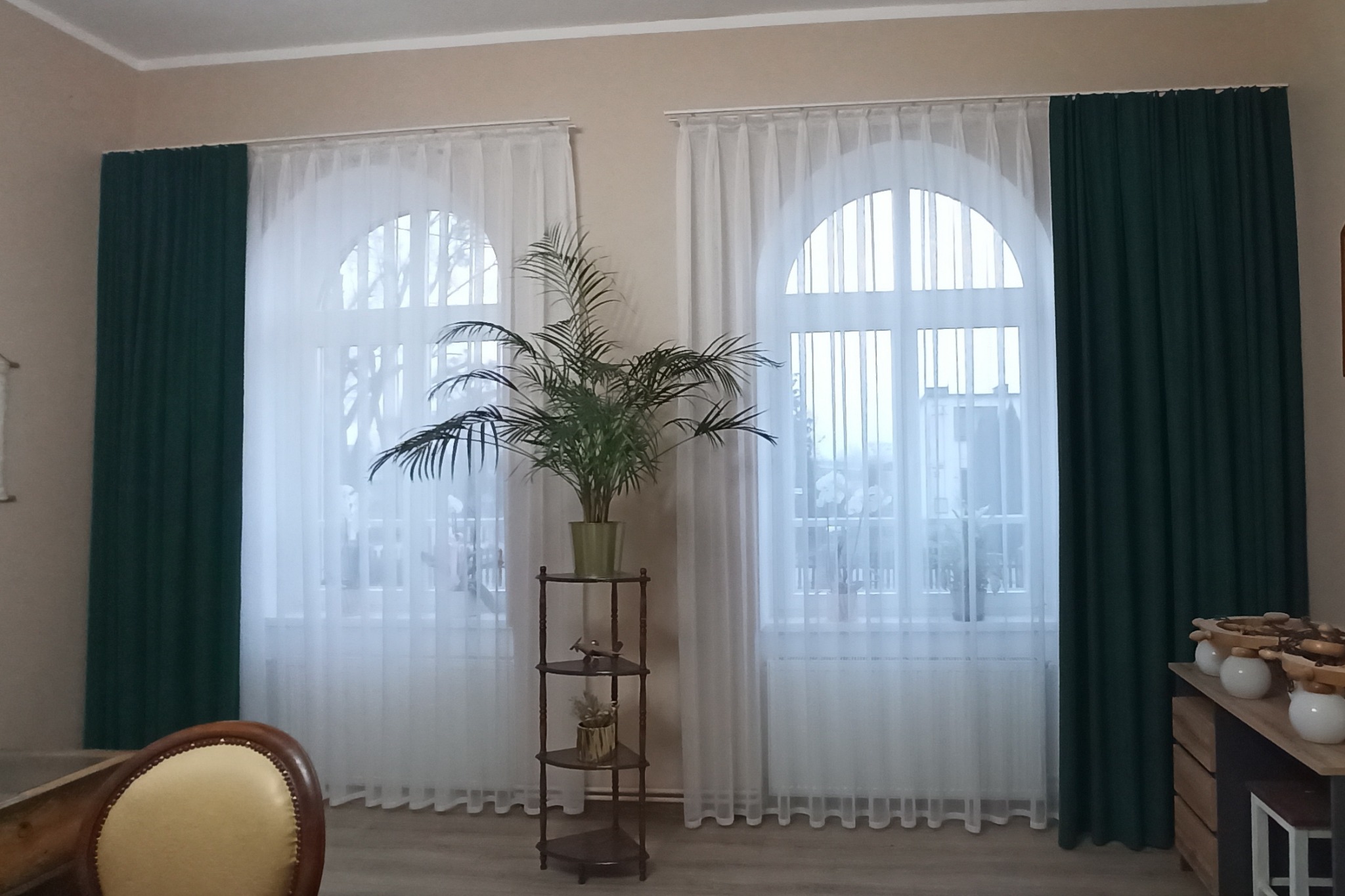 Aranżacja okienna z ciemnozielonymi i białymi, prześwitującymi tkaninami, dopełniona rośliną doniczkową na drewnianym kwietniku i widokiem za oknem.