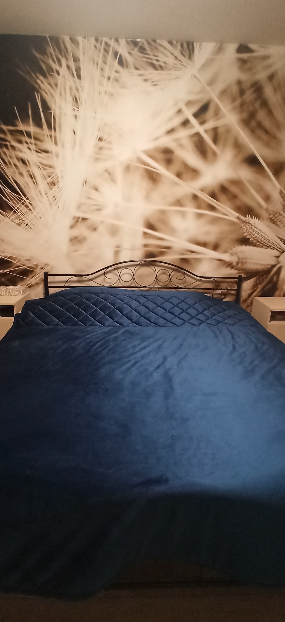 Niebieska, pikowana narzuta na łóżko z metalowym zagłówkiem na tle fototapety z motywem dmuchawca.