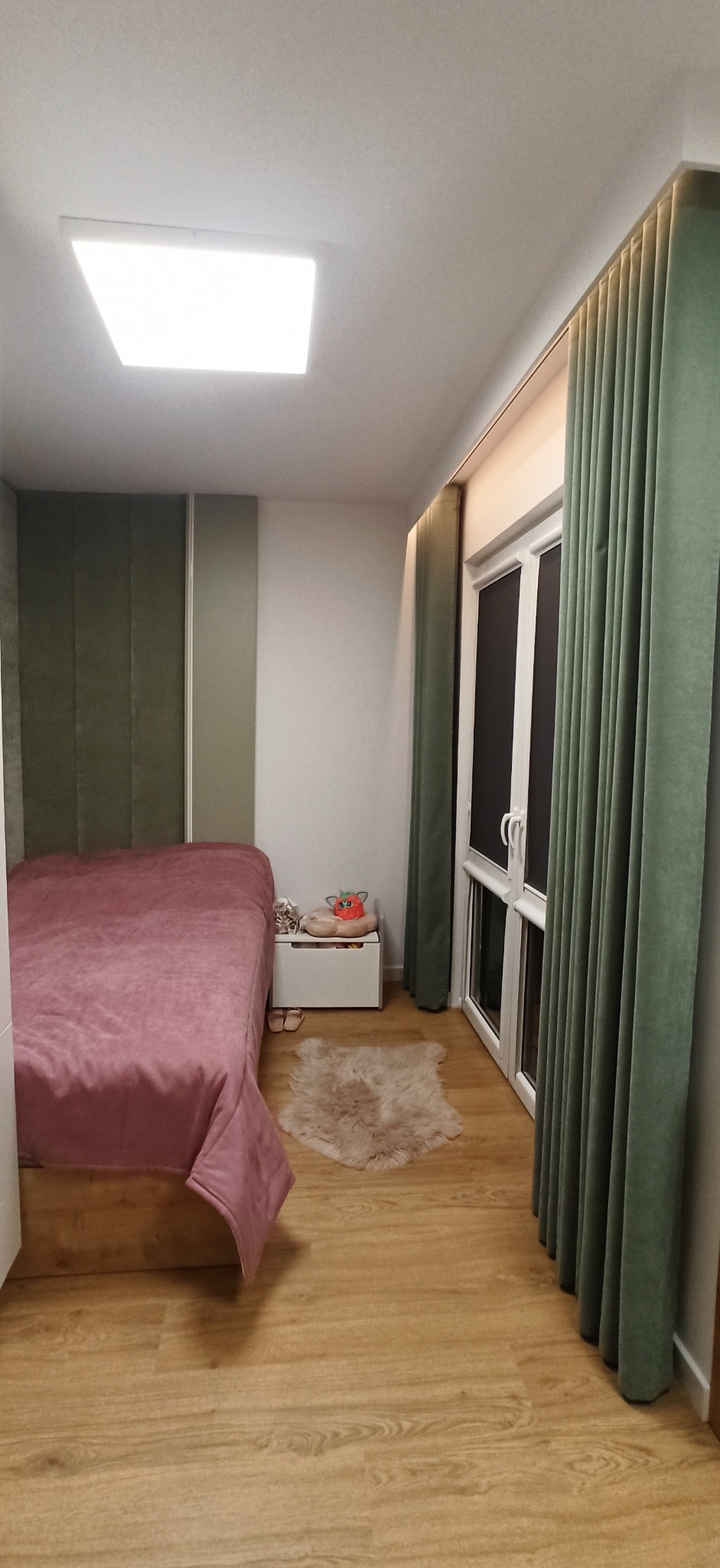 Sypialnia z zasłonami w odcieniach zieleni, różowe łóżko, drewniana podłoga i białe okno.