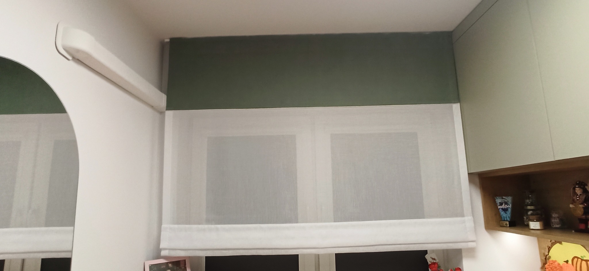 Zasłona rzymska z zielonym pasem górnym, zamontowana w oknie, widok z dołu, obok jasne szafki kuchenne.