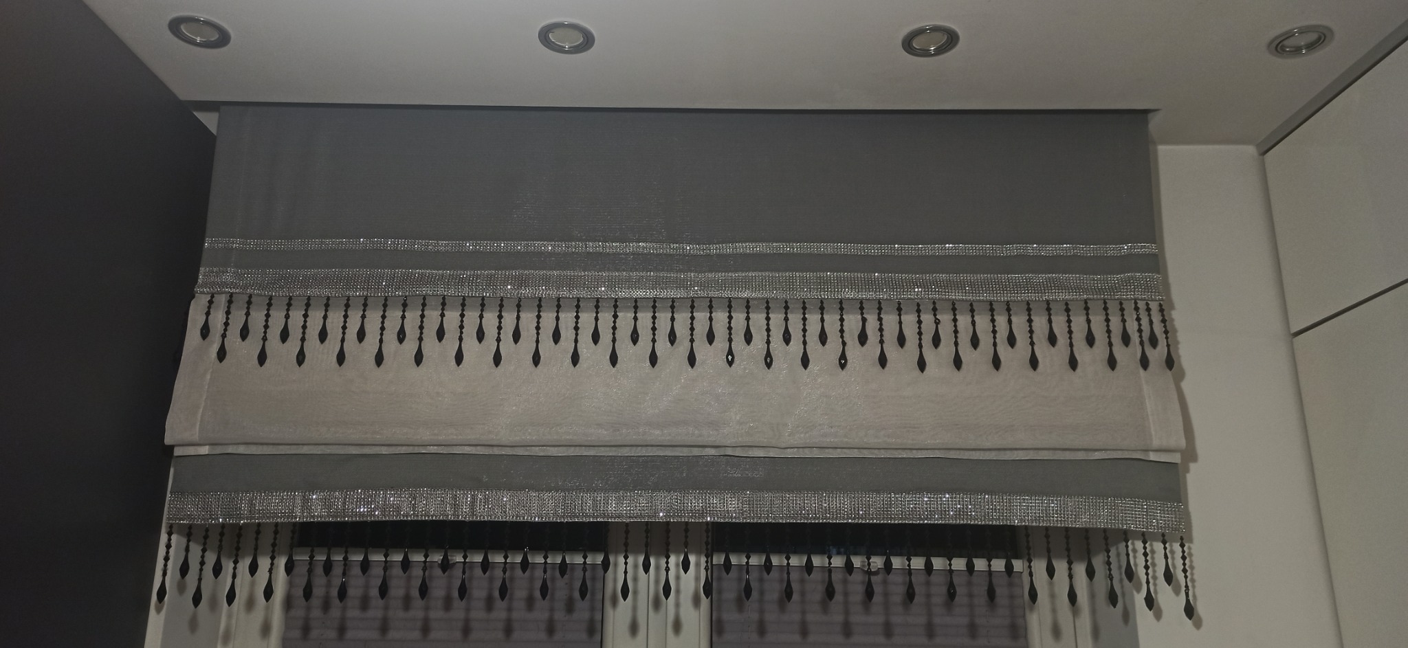 Szara, dwuwarstwowa roleta rzymska z dekoracyjnymi, błyszczącymi pasami i czarnymi, kryształkowymi frędzlami, zamontowana w oknie.