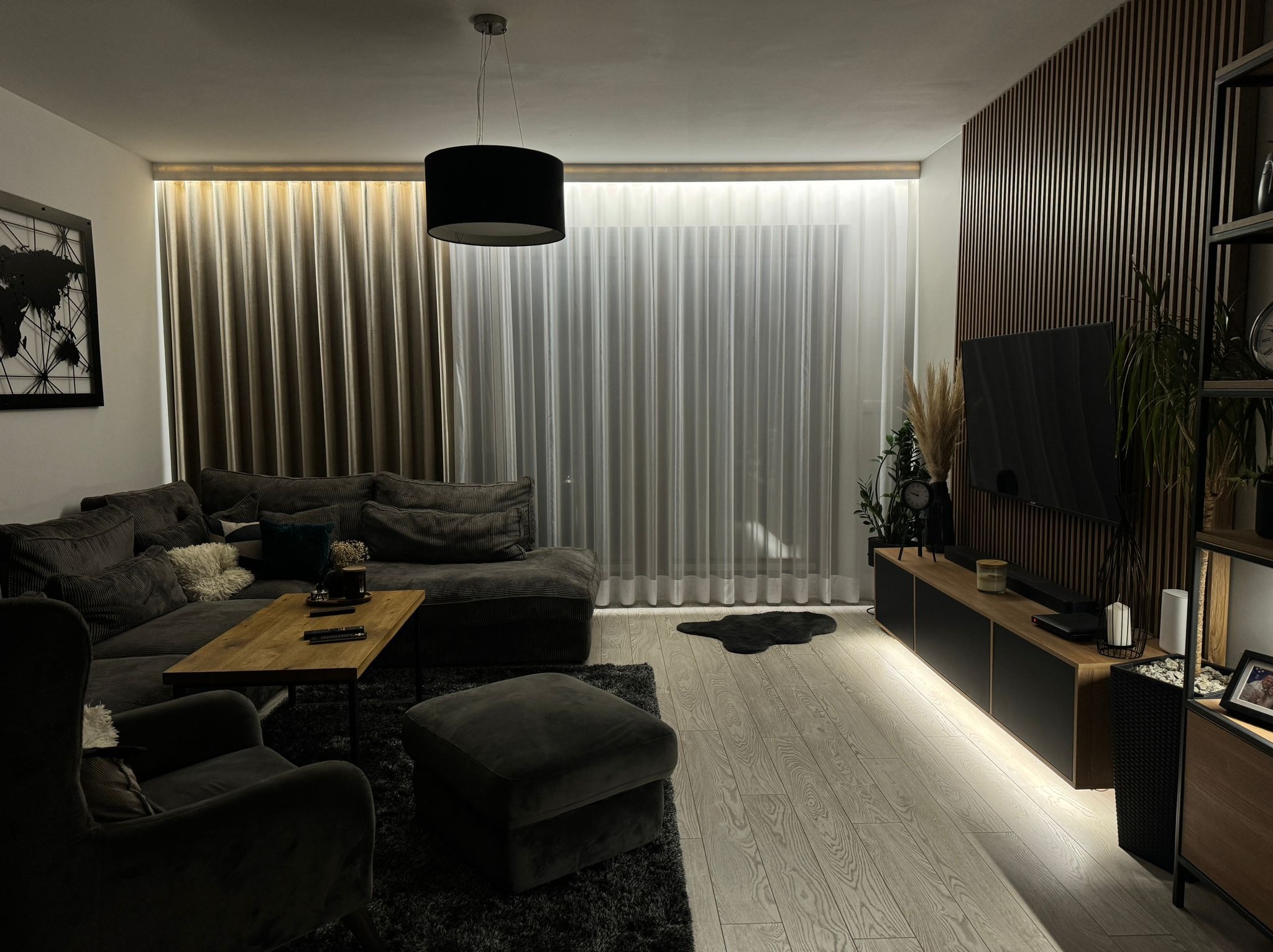 Nowoczesny salon z szarym narożnikiem, drewnianym stolikiem kawowym i zasłonami w odcieniach beżu i bieli, podświetlonymi od góry, tworzącymi miękkie światło.