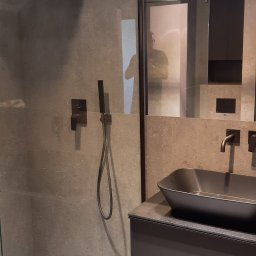 Ultima Art Piotr Majewski - Nowoczesna łazienka z kabiną prysznicową typu walk-in, ciemną armaturą i umywalką nablatową, widoczny również środek czyszczący na podłodze.