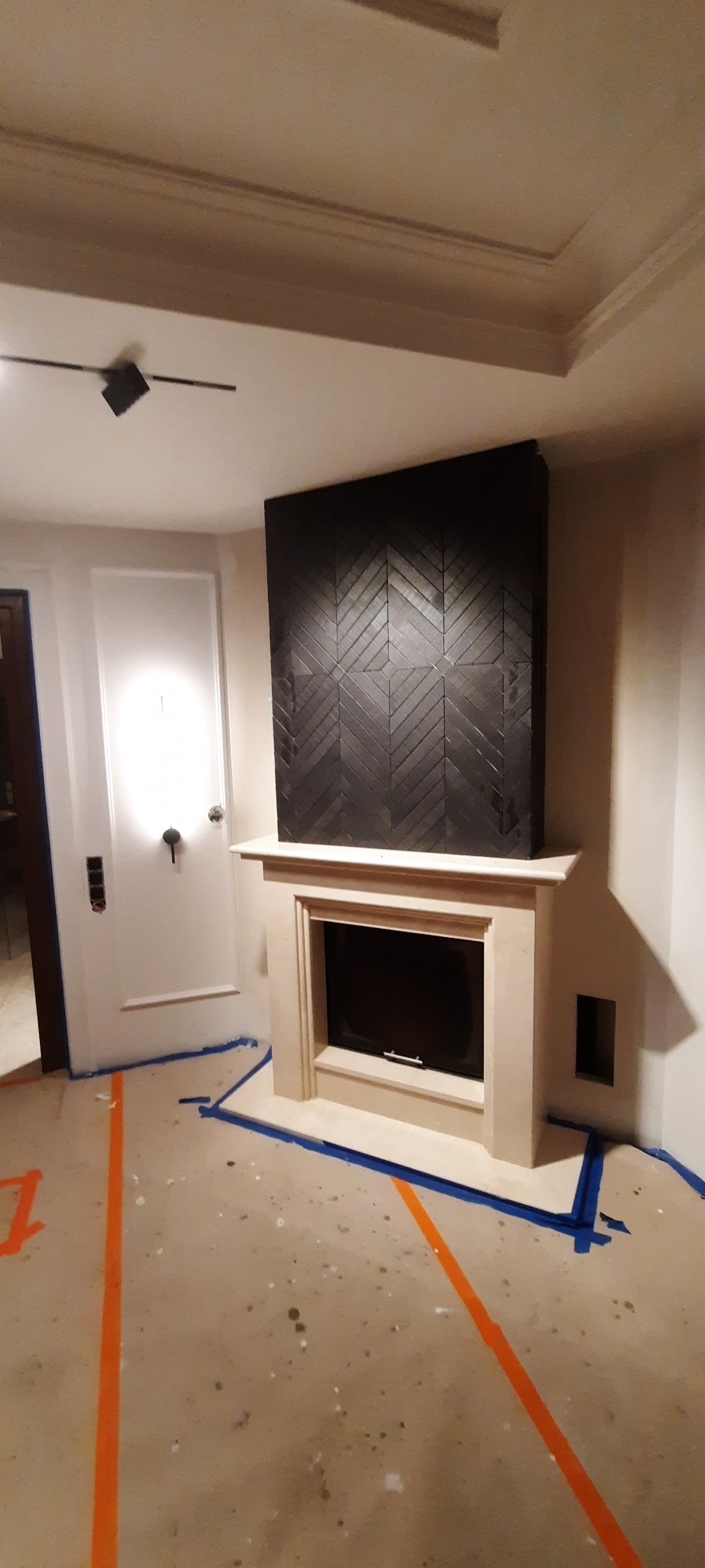 Fragment pokoju z kominkiem obudowanym jasnym kamieniem, nad którym znajduje się ciemny panel z wzorem jodełki; podłoga zabezpieczona taśmą malarską, widoczna lampa sufitowa i białe drzwi...