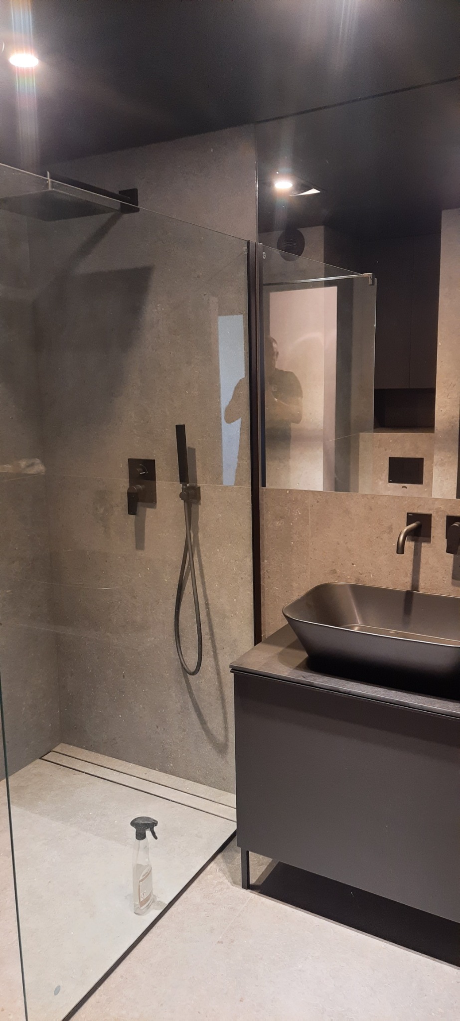 Nowoczesna łazienka z kabiną prysznicową typu walk-in, ciemną armaturą i umywalką nablatową, widoczny również środek czyszczący na podłodze.