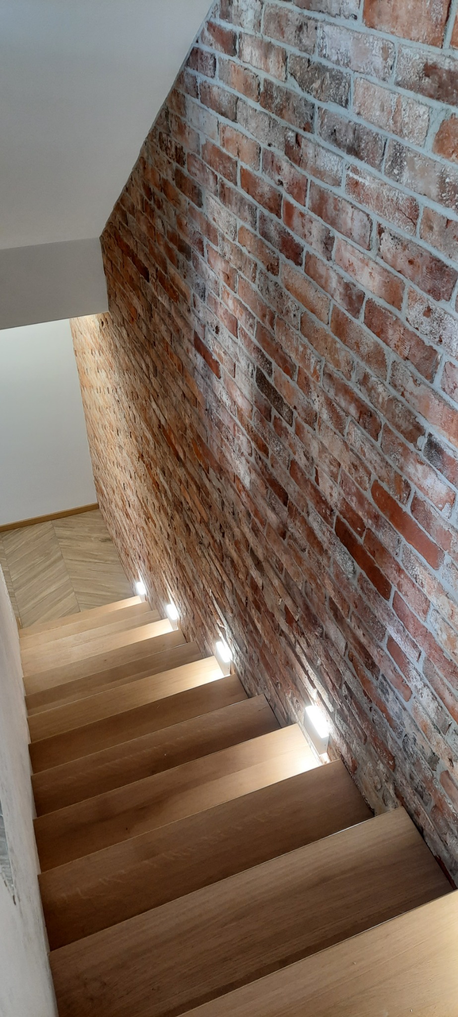Drewniane schody z podświetleniem LED wbudowanym w ścianę z cegły dekoracyjnej, widok z góry.